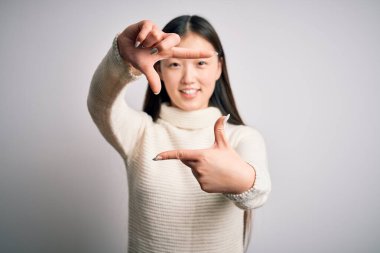 Genç ve güzel Asyalı bir kadın, sıradan bir süveter giyip izole edilmiş arka planda duruyor gülümsüyor ve elleri ve parmakları gülen yüzlü bir çerçeve yapıyor. Yaratıcılık ve fotoğrafçılık kavramı.