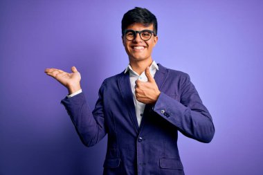 Ceket ve gözlük takan genç yakışıklı iş adamı, mor arka planda avuçlarını gösteriyor ve baş parmaklarıyla hareket ediyor, mutlu ve neşeli gülümsüyor.