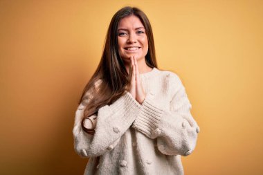 Mavi gözlü genç bir kadın sarı arka planda dikilmiş ellerini birleştirip af diliyor kendinden emin bir şekilde gülümsüyor..