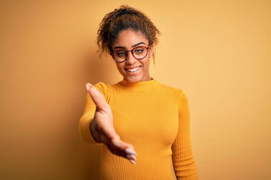 Sarı arka planda süveter ve gözlük takan güzel Afrikalı Amerikalı genç bir kız selamlaşma ve hoş geldin demek için gülümseyerek tokalaşıyor. Başarılı bir iş..