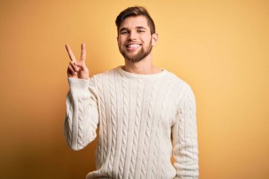 Sakallı, mavi gözlü, sarı arka planda beyaz kazak giyen, gülümseyen ve parmaklarını zafer işareti yapan genç sarışın adam. İki numara..