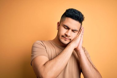 Genç ve yakışıklı bir adam, günlük tişört giyip izole edilmiş sarı arka planda dikiliyor. Yorgun uyuyor, hayal kuruyor ve elleri ile birlikte poz veriyor, gözleri kapalı gülümsüyor..