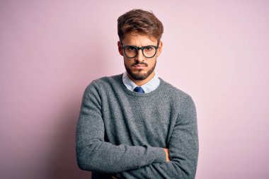 Sakallı, gözlüklü ve süveterli genç bir adam pembe arka plan üzerinde duruyor şüpheci ve gergin, yüzünde çapraz kollu tasvip etmeyen bir ifade. Negatif kişi.