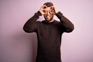 Sakallı ve mavi gözlü genç bir adam pembe arka planda sıradan bir süveter giyiyor. Dürbün gibi jest yapıyor. Dürbün dilini çıkarıyor, gözler parmaklarından bakıyor. Çılgın ifade.