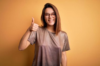 Güzel esmer kız, gündelik tişört ve gözlük takıp izole edilmiş sarı arka planda mutlu bir el hareketi yapıyor. Kameraya bakıp başarıyı gösteren ifade onaylanıyor.