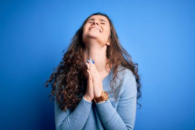 Kıvırcık saçlı, mavi süveter giyen, izole edilmiş arka planda el ele yalvaran ve dua eden, yüzünde umut ifadesi olan çok duygusal ve endişeli genç bir kadın. Yalvarıyorum..