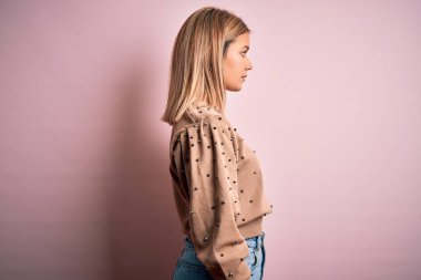 Pembe izole edilmiş arka planda moda süveteri giyen genç, güzel sarışın kadın yan yana bakıyor, rahat poz veriyor, kendine güvenen bir gülümsemesi var..