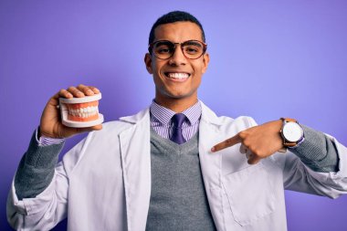 Genç, Afro-Amerikan dişçi, mor arka planda takma dişleri olan, kendini işaret eden sürpriz yüzlü,