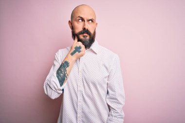 Sakallı, zarif tişörtlü, soyutlanmış pembe arka planda eli çenesinde soru sormayı düşünen yakışıklı kel bir adam. Düşünceli bir ifade. Düşünceli bir yüzle gülümsüyor. Şüphe kavramı.