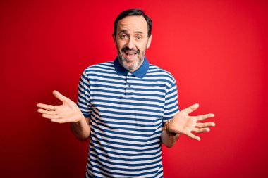 İzole edilmiş kırmızı arka planda dikilen sıradan çizgili polo giyen, kolları ve elleri havada, kafası karışmış orta yaşlı, ağarmış bir adam. Şüphe kavramı.