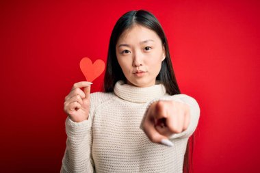 Genç Asyalı bir kadın kırmızı kalp kâğıdı şeklinde romantik bir kağıt tutarak izole edilmiş arkaplanı işaret ederek kamerayı işaret ediyor ve sen, el işareti, ön taraftan pozitif ve kendinden emin bir hareket.