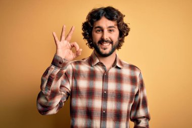 Sakallı, sıradan tişörtlü, sarı arka planda duran yakışıklı genç adam el ve parmaklarıyla 