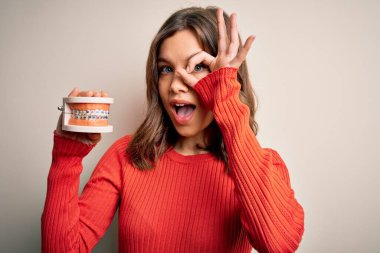 Sarışın genç kız diş protezini izole edilmiş arka planda tutuyor. Gülümsüyor, gülümsüyor. El ve göz işaretleriyle parmaklarından bakıyor.