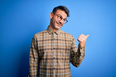 Genç ve yakışıklı bir adam, günlük gömlek ve gözlük takıyor mavi arka planda gülümsüyor, gülümsüyor ve başparmağıyla yan tarafa bakıyor..