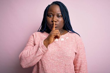 Siyah saçlı, uzun saçlı, pembe arka planda sıradan süveter giyen Afro-Amerikan kadın sessiz olup dudaklarını öpmek istiyor. Sessizlik ve gizli konsept.