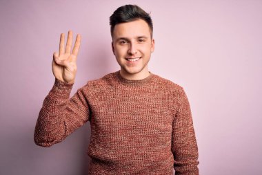 Pembe izole edilmiş arka plan üzerine sıradan bir kazak giyen yakışıklı beyaz adam üç numarayı işaret ederken kendinden emin ve mutlu bir şekilde gülümsüyor..