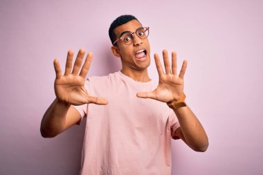 Yakışıklı Afro-Amerikalı adam pembe arka plan üzerinde günlük tişört ve gözlük takıyor korku ifadesinden korkuyor ve dehşete düşüyor. El hareketlerini bırak, şok içinde bağır. Panik konsepti.
