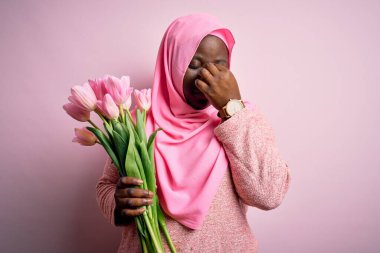 Genç, Afro-Amerikan, iri yarı, Müslüman bir kadın elinde pembe lale buketi, burnu ve gözleri sürtünmekten yorgun düşmüş ve başı ağrıyormuş. Stres ve hüsran kavramı.