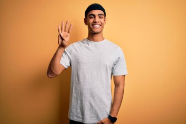 Yakışıklı Afro-Amerikan sporcu sarı arka planda spor giysisi giyiyor ve parmaklarıyla dört numarayı gösterirken kendinden emin ve mutlu gülümsüyor..