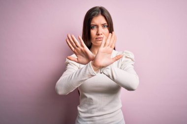 Mavi gözlü genç bir kadın pembe arka planda günlük beyaz tişört giyiyor. Reddedilmiş yüz ifadesi kollarını kavuşturuyor. Negatif işaret veriyor, kızgın yüz ifadesi takınıyor.