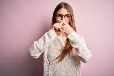 Genç, güzel, kızıl saçlı bir kadın pembe arka planda gözlük ve süveter takıyor. Reddedilme ifadesi olumsuz işaret veriyor.
