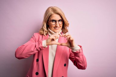 Orta yaşlı, güzel, sarışın, pembe ceketli ve gözlüklü bir iş kadını. Aşkla gülümseyen, kalp sembolü ve elleri olan bir kadın. Romantik konsept.