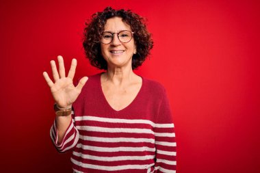 Orta yaşlı güzel kıvırcık saçlı kadın kırmızı arka planda sıradan çizgili süveter giyiyor ve parmaklarını 5 numarayı gösterirken kendinden emin ve mutlu bir şekilde gülümsüyor..