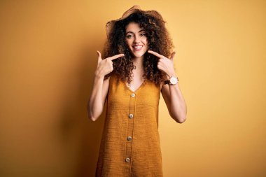 Güzel esmer kadın tatilde. Kıvırcık saçlı, piercing takan, şapkalı, elbiseli, neşeli gülümseyen, parmakları, dişleri ve ağzıyla işaret eden. Diş sağlığı konsepti.