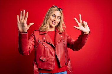 Genç, sarışın, sıradan bir ceket giyen, kırmızı arka plan üzerinde duran ve 8 numaralı parmaklarıyla işaret eden, kendinden emin ve mutlu bir şekilde gülümseyen bir kadın..