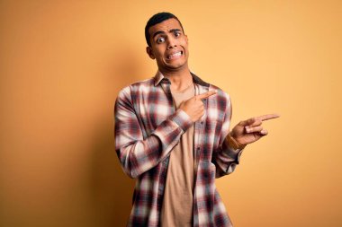 Sarı arka planda dikilen rahat gömlekli, genç ve yakışıklı Afro-Amerikalı bir adam iki eliyle endişeli ve endişeli bir ifadeye işaret ediyor.