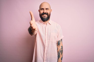 Sakallı ve dövmeli yakışıklı, pembe arka planda rahat gömleği olan yakışıklı, dostça gülümseyerek selamlaşan ve hoş geldin diyen bir adam. Başarılı bir iş..