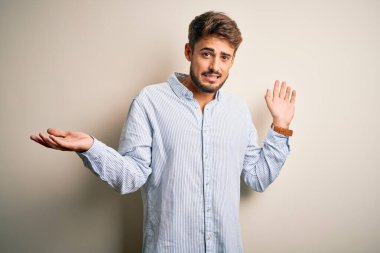 Beyaz arka plan üzerinde duran çizgili tişörtlü, sakallı, genç ve yakışıklı bir adam. Kolları ve elleri havada, kafası karışmış bir ifade. Şüphe kavramı.