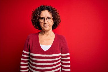 Orta yaşlı güzel kıvırcık saçlı kadın kırmızı arka planda sıradan çizgili süveter giyiyor şüpheci ve gergin, sorun yüzünden surat asıyor. Negatif kişi.