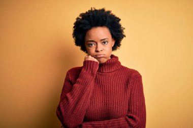 Kıvırcık saçlı, balıkçı yaka kazak giyen genç, güzel Afro-Amerikan kadın kollarını kavuşturup depresyondan sıkılmış ve yorgun göründüğünü düşünüyor..