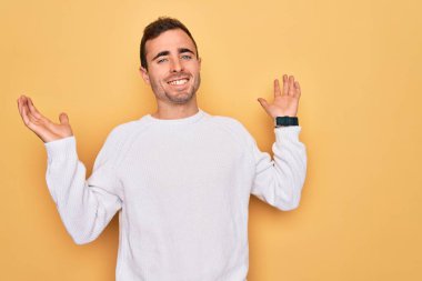 Mavi gözlü, sarı arka planda dikilen sıradan süveter giyen, elleri ve kolları havada, kafası karışmış genç, yakışıklı bir adam. Şüphe kavramı.