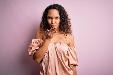 Kıvırcık saçlı, rahat tişörtlü, pembe arka planda dikilmiş kameraya bakıp havada öpücükler saçan, güzel ve seksi genç bir kadın. Aşk ifadesi.