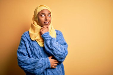 Genç Afrikalı Afro kadın, izole edilmiş sarı arka planda Müslüman başörtüsü takıyor. Elleri çenesinde soru sorarken, düşünceli bir ifade takınıyor. Düşünceli bir yüzle gülümsüyor. Şüphe kavramı.