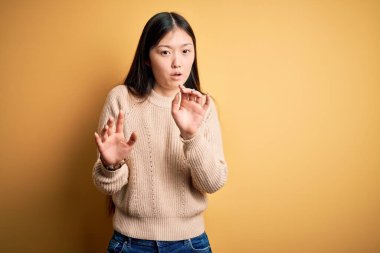 Sarı arka plan üzerine sıradan süveter giyen genç, güzel Asyalı bir kadın. Avuçlarını kaldırıyor, reddediyor ve iğrenç bir ifadeyle reddediyor. Dur ve yasakla.