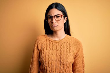 Sarı arka plan üzerine gözlük takan genç ve güzel İspanyol kadın şüpheci ve gergin, sorun yüzünden surat asıyor. Negatif kişi.