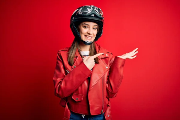若い美しいブルネットのオートバイのヘルメットと赤いジャケットを身に着けているオートバイの女性は驚いて 手で提示しながら 指で指してカメラに笑みを浮かべて — ストック写真