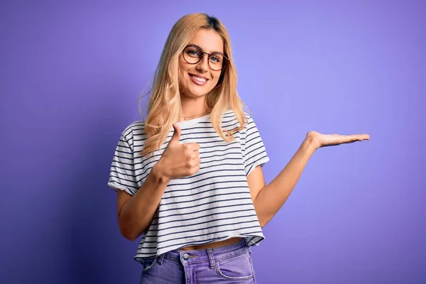 Young beautiful blonde woman wearing striped t-shirt and glasses over purple background Showing palm hand and doing ok gesture with thumbs up, smiling happy and cheerful