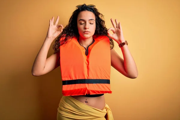 Young beautiful woman with curly hair wearing orange lifejacket over yellow background relax and smiling with eyes closed doing meditation gesture with fingers. Yoga concept.