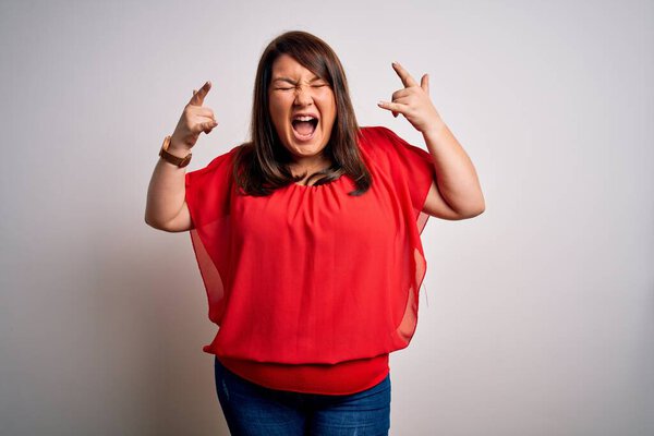 Beautiful brunette plus size woman wearing casual red t-shirt over isolated white background shouting with crazy expression doing rock symbol with hands up. Music star. Heavy music concept.