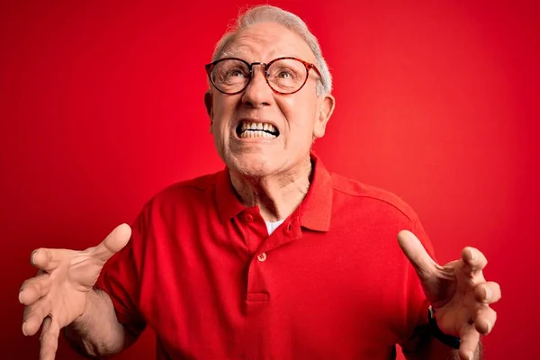 Grey haired senior man wearing glasses and casual t-shirt over red background crazy and mad shouting and yelling with aggressive expression and arms raised. Frustration concept.