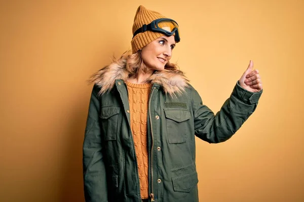 中年漂亮的金发滑雪者女人穿着雪地运动鞋和滑雪护目镜看起来很自豪 微笑着向旁边竖起大拇指 — 图库照片