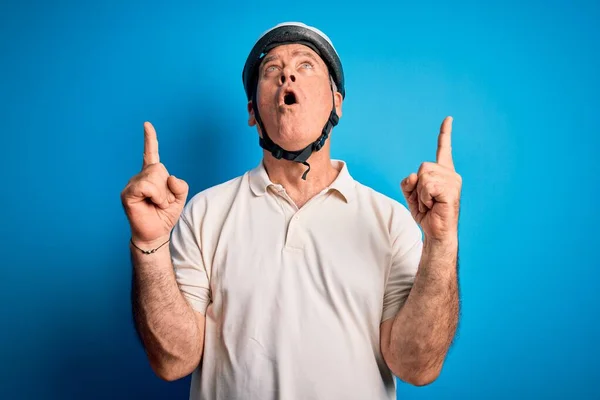 Middle age hoary cyclist man wearing bike security helmet over isolated blue background amazed and surprised looking up and pointing with fingers and raised arms.