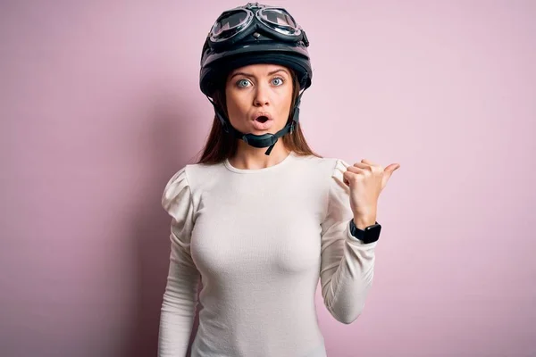 若いです美しいですオートバイの女性とともに青い目ピンクの背景の上に元ヘルメットを身に着けています側面に手指で指摘し 口を開けて驚くような式 — ストック写真