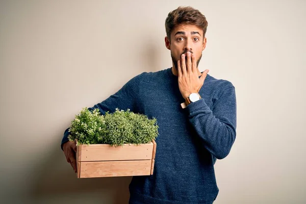 Young gardener man with beard holding box with plants standing over white background cover mouth with hand shocked with shame for mistake, expression of fear, scared in silence, secret concept