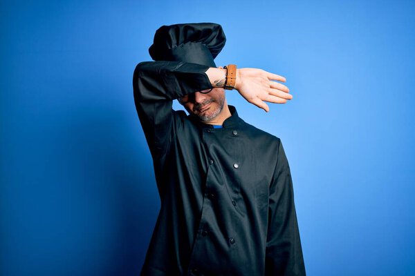 Young handsome chef man wearing cooker uniform and hat over isolated blue background covering eyes with arm, looking serious and sad. Sightless, hiding and rejection concept