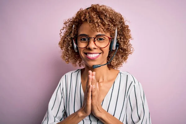 African american curly call center agent woman working using headset over pink background praying with hands together asking for forgiveness smiling confident.